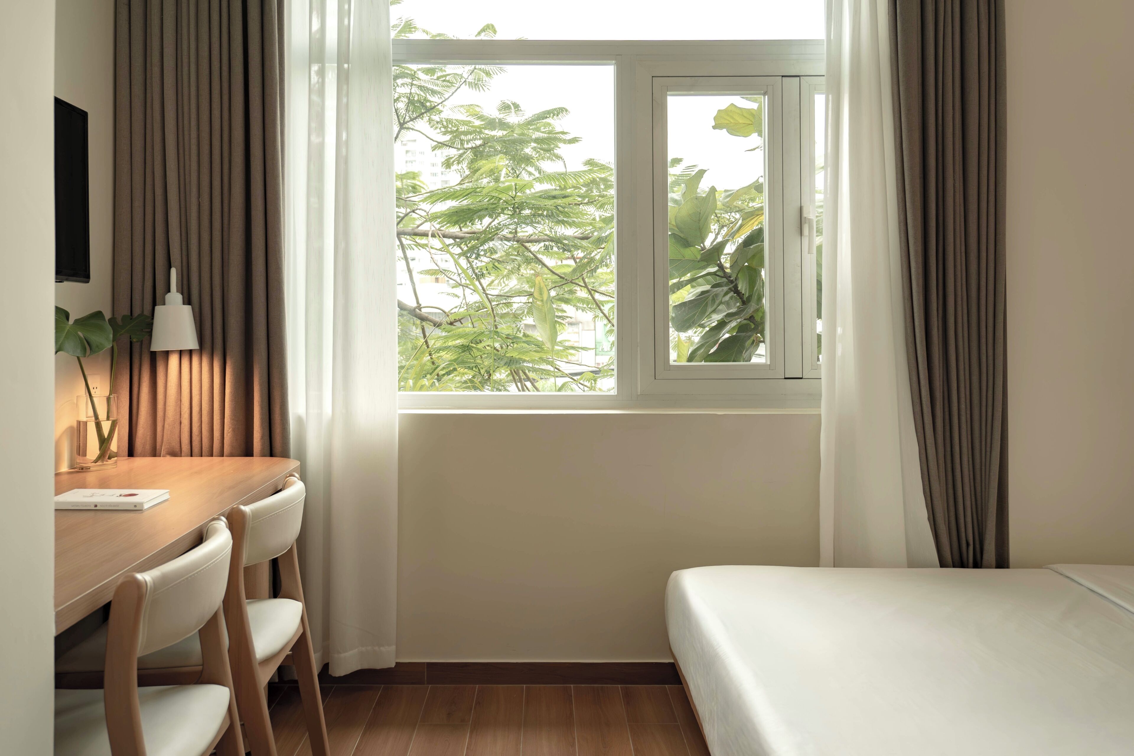 A room with a bed, a desk with chairs, a window with curtains, and a plant.
