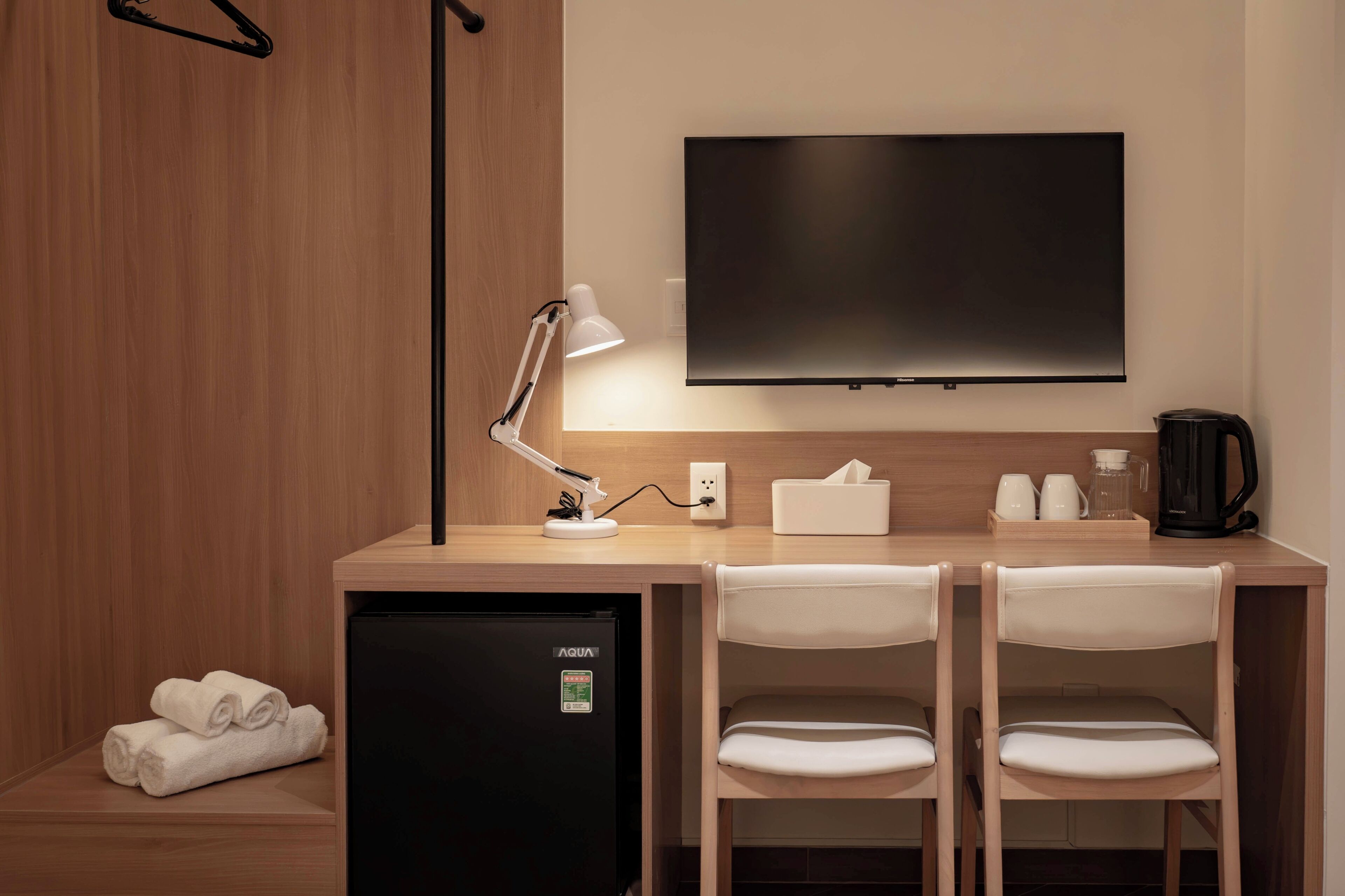 A hotel room with a desk, two chairs, a flat-screen TV, a desk lamp, a mini-fridge, a coffee maker, and a towel on the floor.