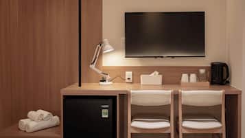 Ein Hotelzimmer mit Schreibtisch, zwei Stühlen, einem Flachbildfernseher, einer Schreibtischlampe, einem Minikühlschrank, einer Kaffeemaschine und einem Handtuch auf dem Boden.