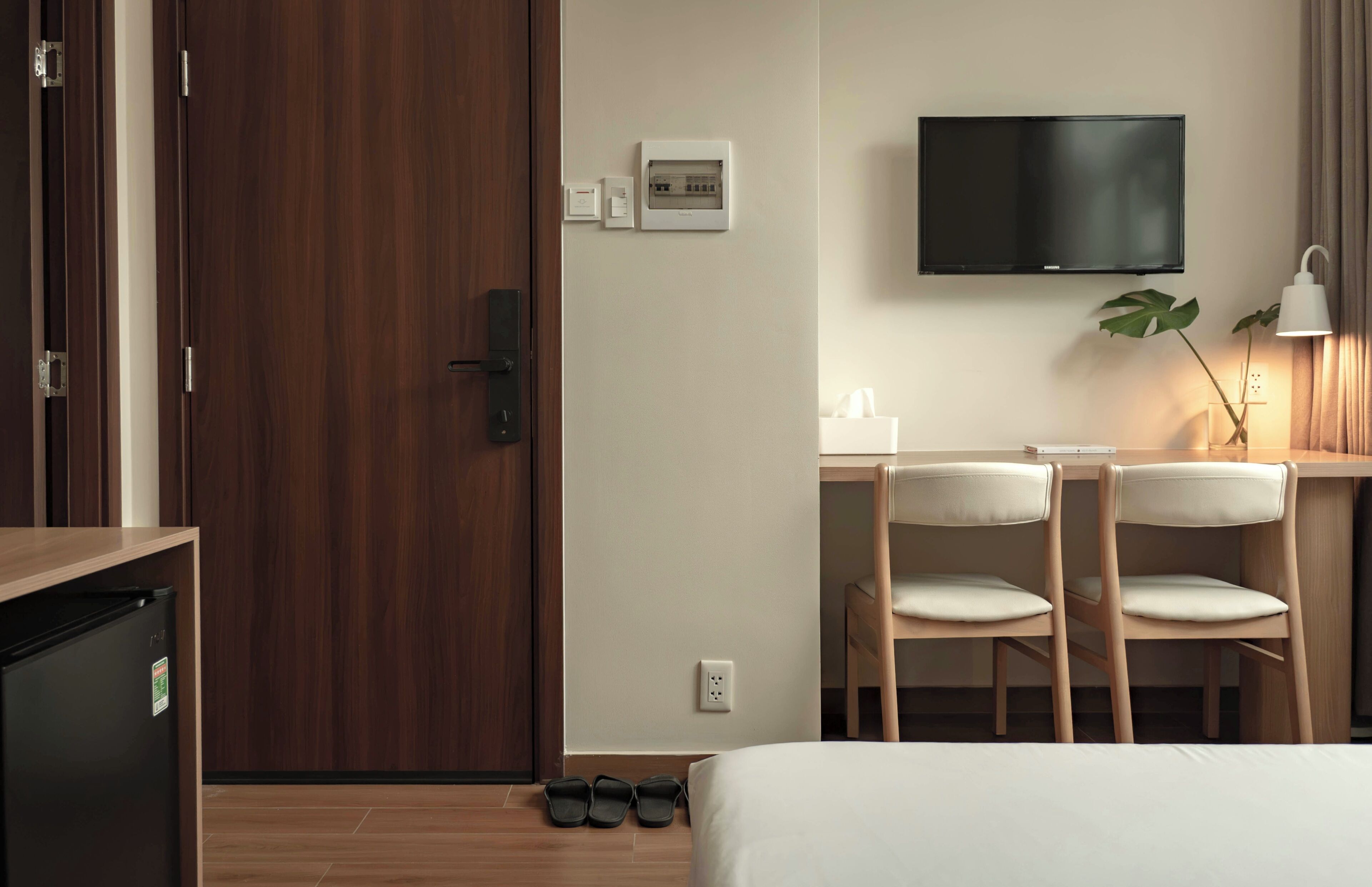 A hotel room with a wooden door, a desk with two chairs, a television, a bedside table, a lamp, and a bed.