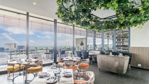 A rooftop restaurant with tables and chairs, a bar area, and a large window offering a city view. - One Rosebank Apartment- Studio (Johannesburg)