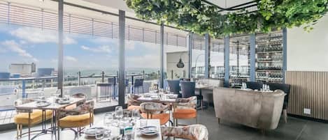 Um restaurante no terraço com mesas, cadeiras, um bar e uma grande janela com vista para a cidade.