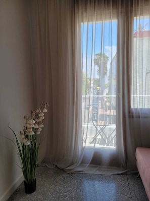 Une pièce dotée d’une grande fenêtre ornée de rideaux légers, d’une plante en pot aux fleurs blanches et offrant une vue sur un balcon aménagé avec du mobilier d’extérieur.