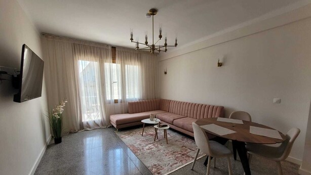 Un soggiorno con un divano, un tavolo da pranzo con sedie, un lampadario e una TV a parete.