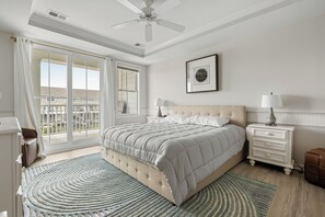 Un dormitorio con una cama grande, un ventilador de techo, una ventana con cortinas, una mesita de noche con lámpara y un cuadro en la pared.