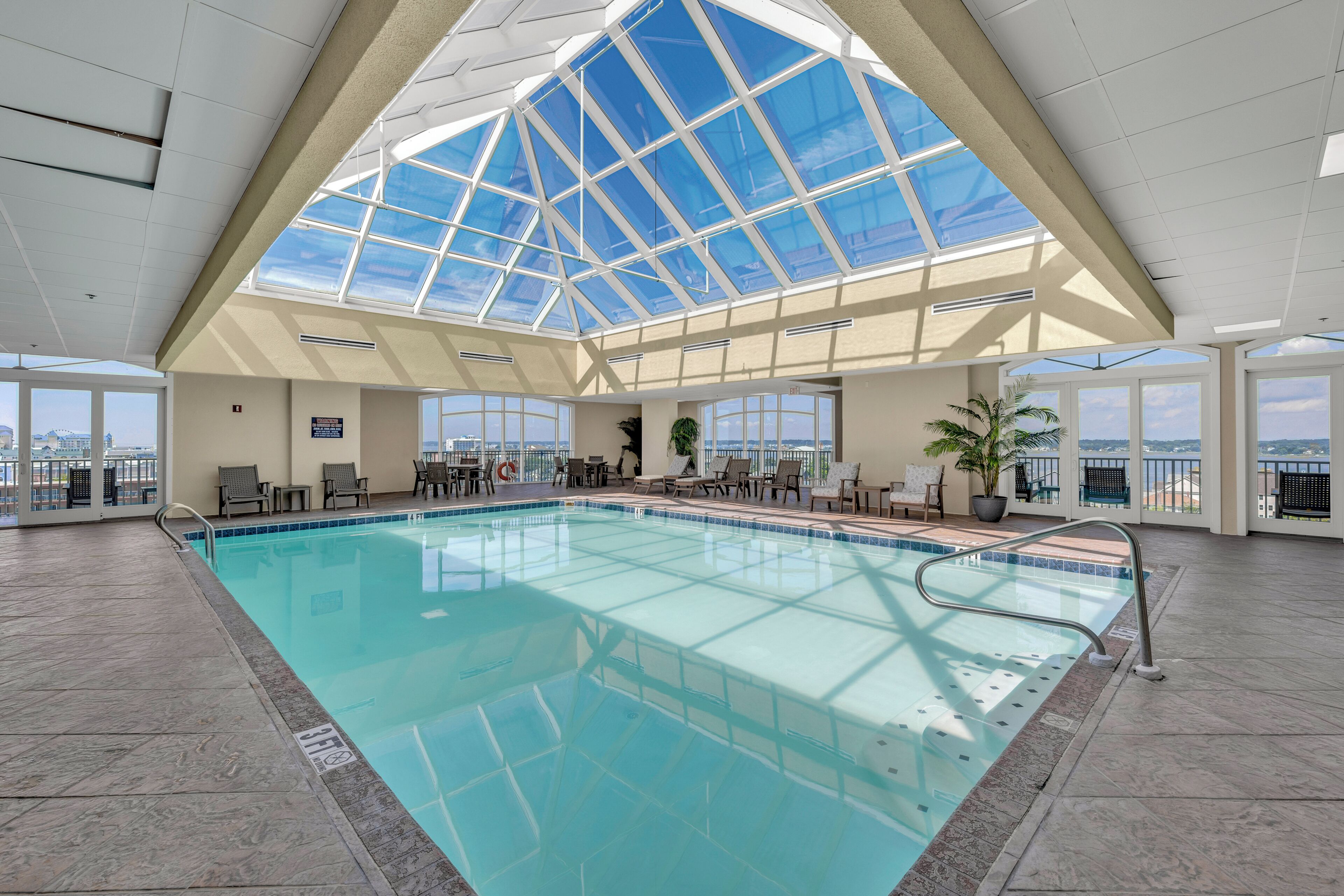 An indoor swimming pool with a glass ceiling, lounge chairs, and a view of the city.