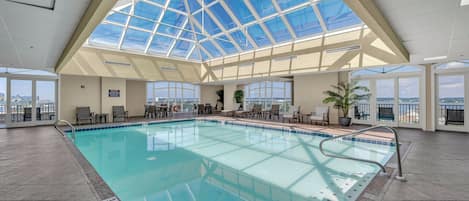 An indoor swimming pool with a glass ceiling, lounge chairs, and a view of the city.