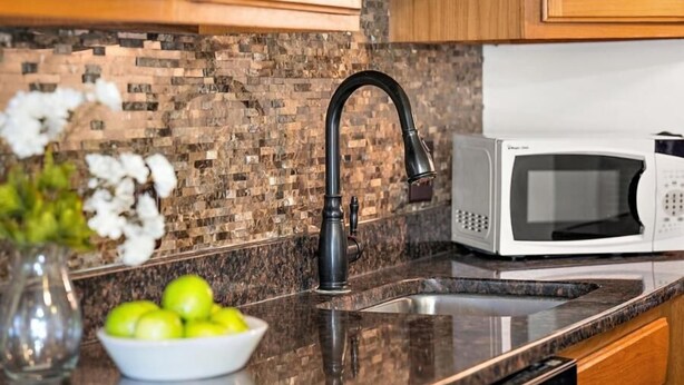 A kitchen with a granite countertop, a stainless steel sink, a white microwave, and wooden cabinets. - Jamaica Day Break N 407 – Bayside Comfort in the Heart of Midtown (Ocean City)