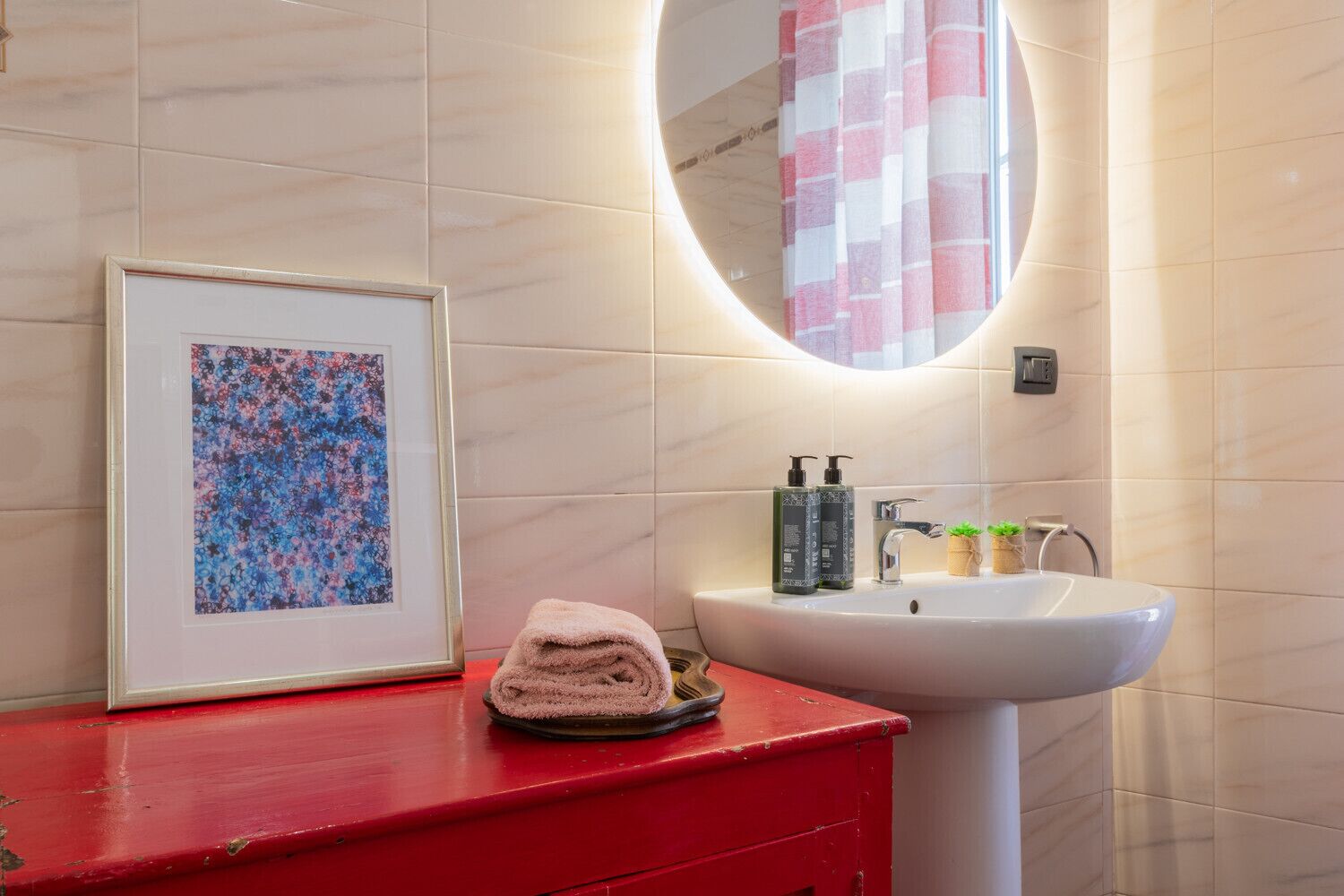 Ein Badezimmer mit rotem Waschtisch, einem floralen Bild in einem Rahmen, einem runden Spiegel und einem Waschbecken mit Toilettenartikeln.