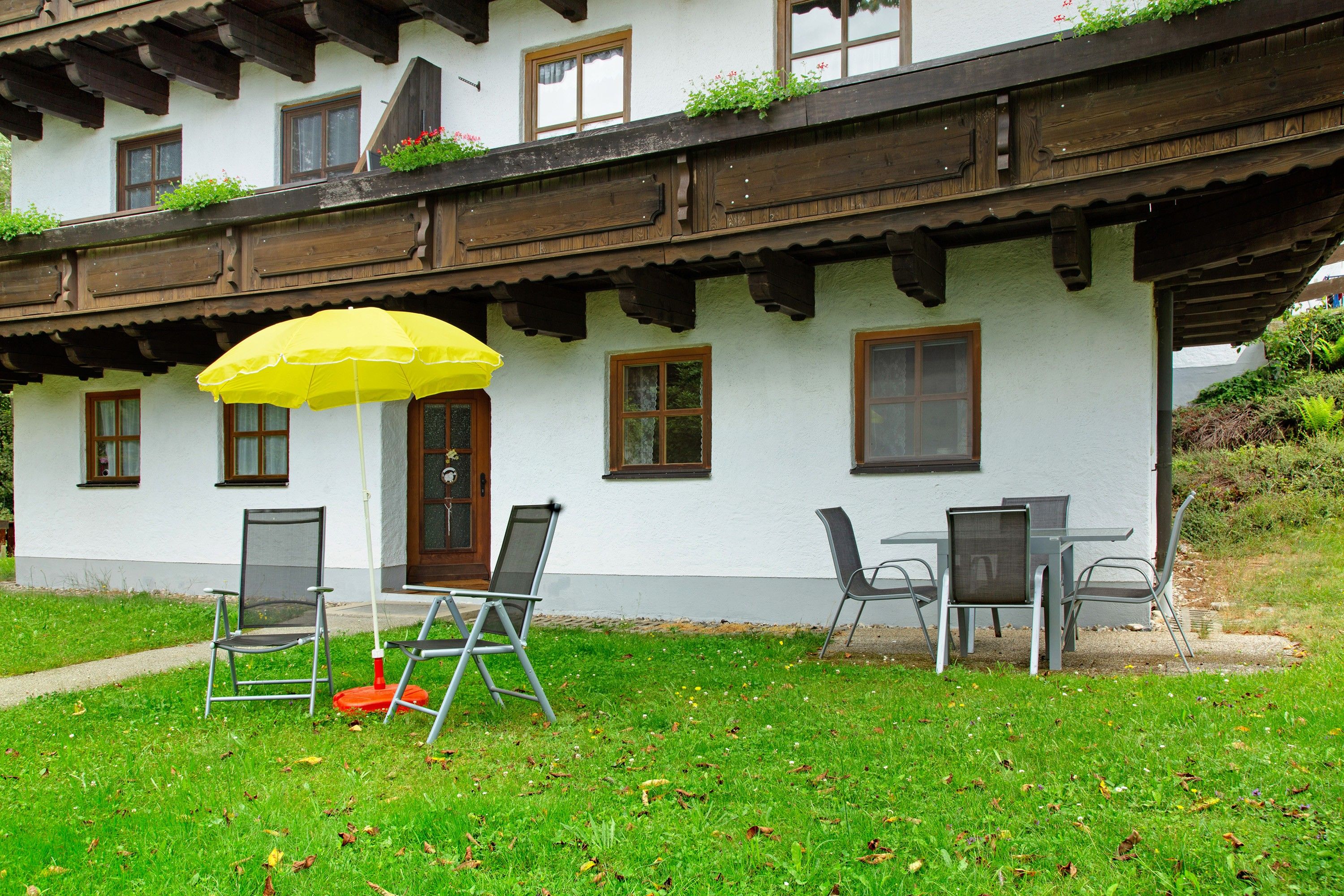 Un bâtiment blanc avec des balcons en bois, une porte et des fenêtres. On peut voir deux chaises et un parapluie jaune à l’extérieur.