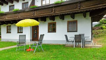 Un bâtiment blanc avec des balcons en bois, une porte et des fenêtres. On peut voir deux chaises et un parapluie jaune à l’extérieur.