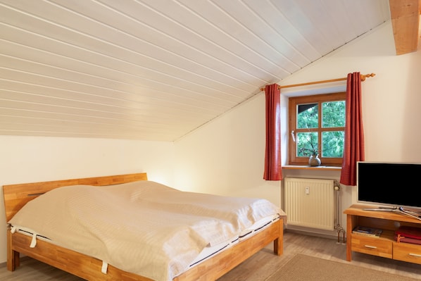 Un dormitorio con cama de madera, ventana con cortinas, radiador y televisor sobre un mueble de madera.