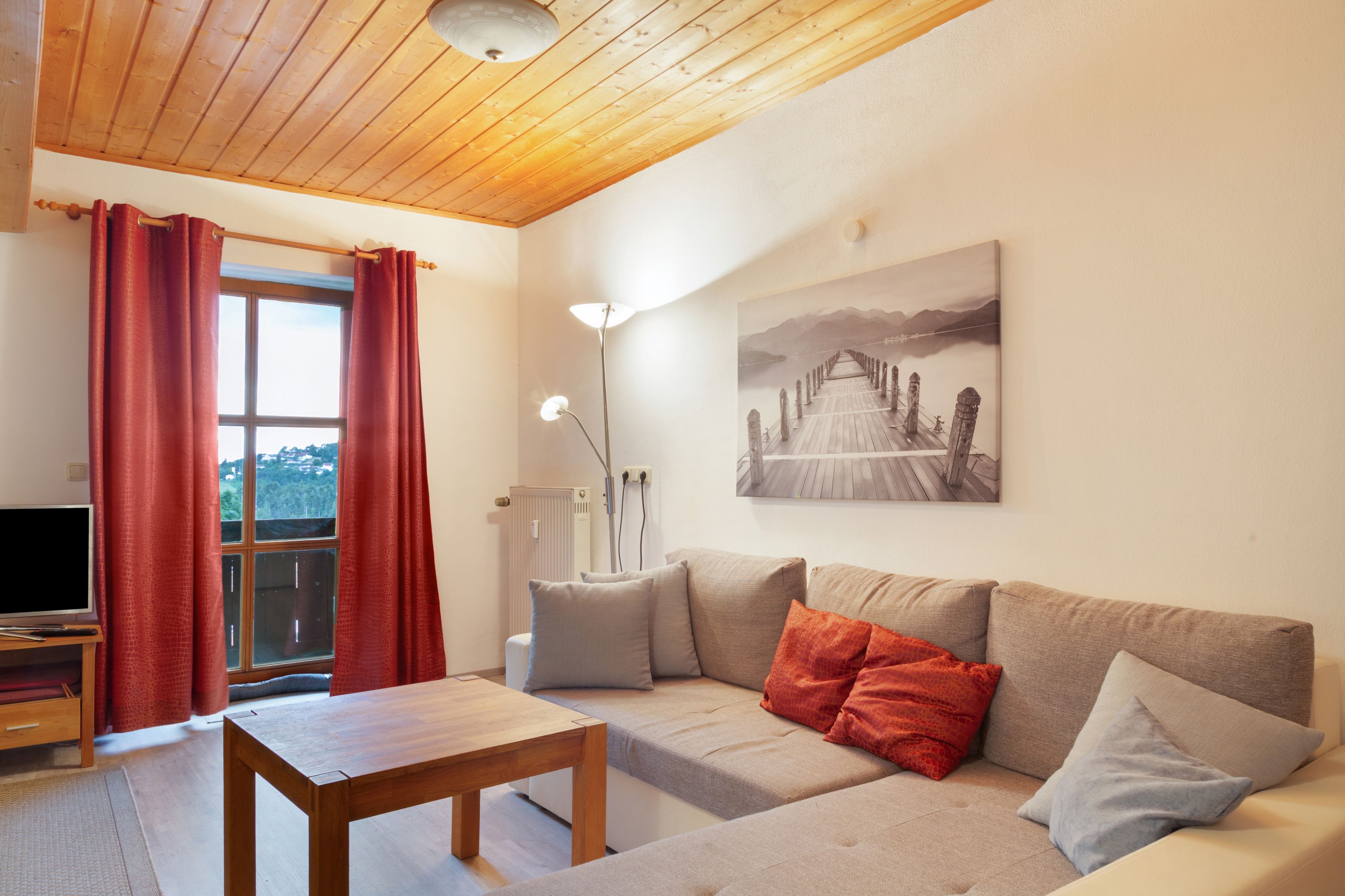 A living room with a wooden ceiling, a sofa with cushions, a table, a floor lamp, and a painting on the wall.