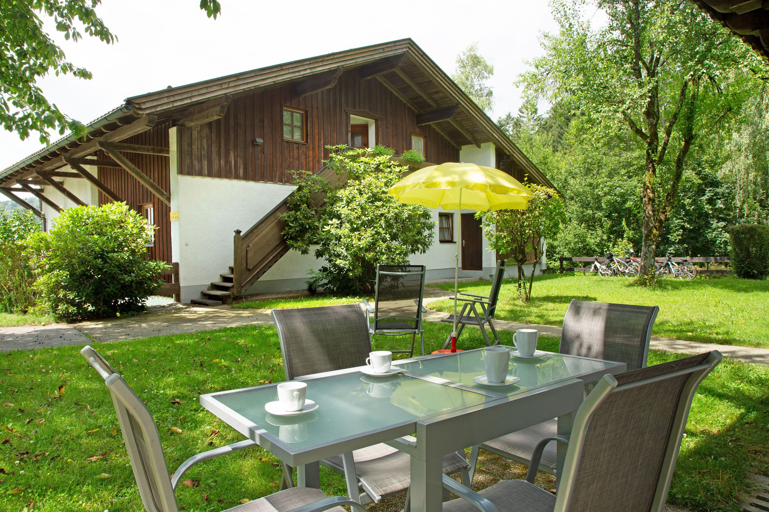 Een glazen tafel met stoelen en een gele parasol in een tuin.