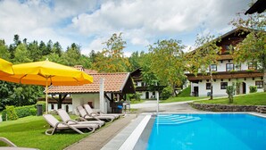 A swimming pool with lounge chairs and umbrellas, surrounded by greenery and a building with a sloped roof. - Apartment Hauzenberg with Indoor & Outdoor Pool, Sauna, Lake View (Hauzenberg)