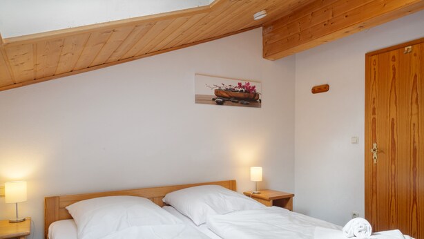A bedroom with a wooden ceiling, a bed with white bedding, two bedside lamps, and a wooden door. - Apartment Hauzenberg with Indoor & Outdoor Pool, Sauna, Lake View (Hauzenberg)