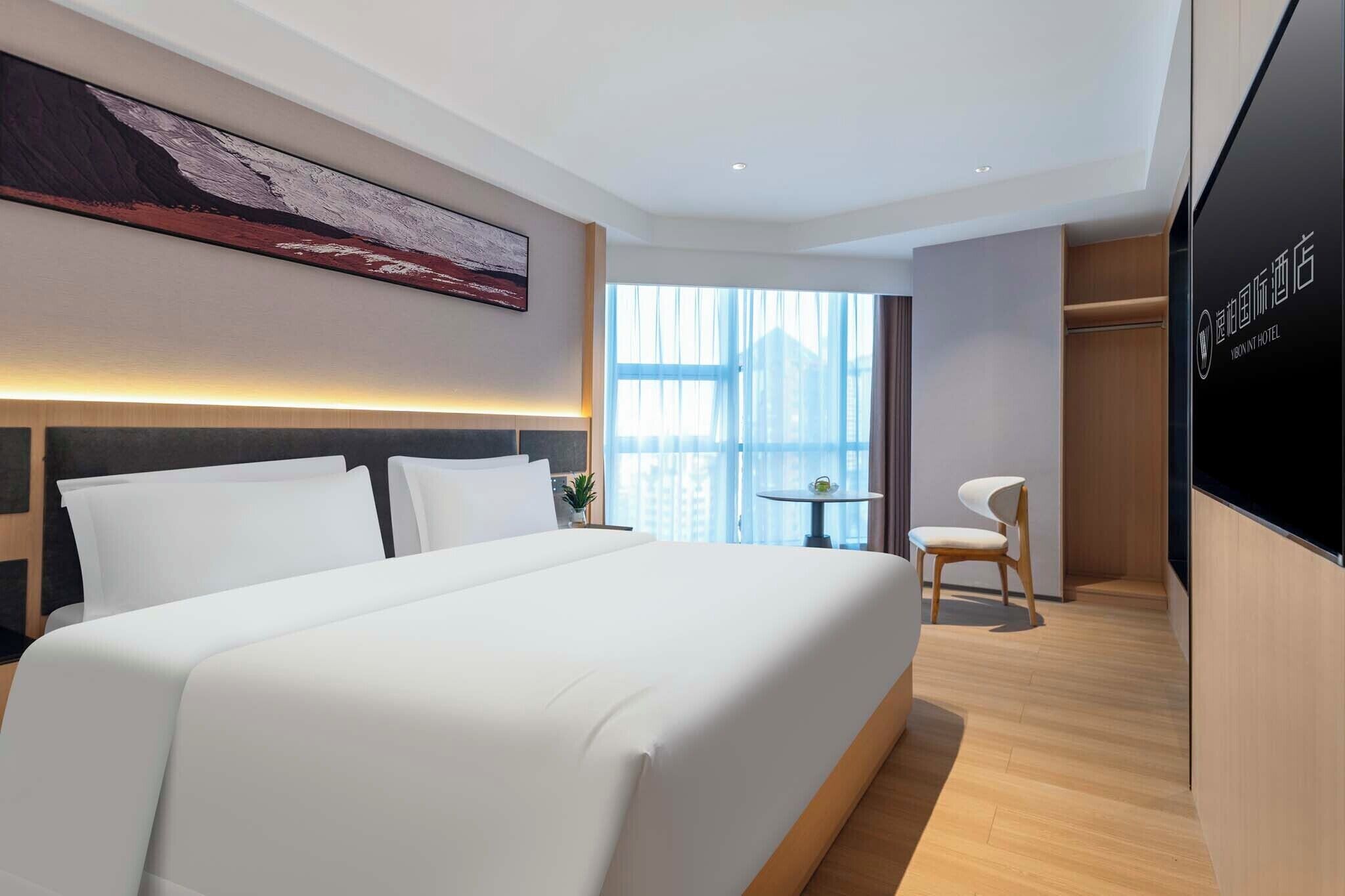 A hotel room with a large bed, a desk, and a chair. There is a window with a view of buildings.
