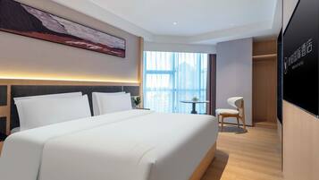 A hotel room with a large bed, a desk, and a chair. There is a window with a view of buildings.