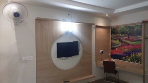 Een kamer met een wandgemonteerde televisie, een ventilator, een bureau met stoel en een groot raam met een schilderij.