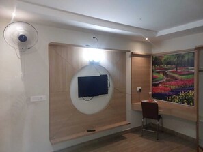 Une pièce équipée d’un téléviseur fixé au mur, d’un ventilateur sur pied, d’un bureau avec une chaise et d’une grande fenêtre ornée d’un tableau.