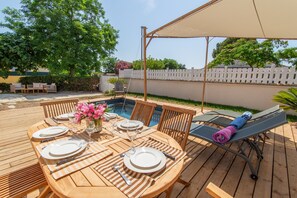 Eine Holzterrasse mit Pooltisch, eingerichtet zum Essen, einem Liegestuhl und einem Pool mit weißem Zaun im Hintergrund.