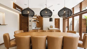 A modern dining area with a round table and six chairs, three pendant lights, and a kitchen visible through an open door.