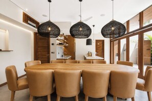 A modern dining area with a round table and six chairs, three pendant lights, and a kitchen visible through an open door. - Villa NA by GORO (Badung)