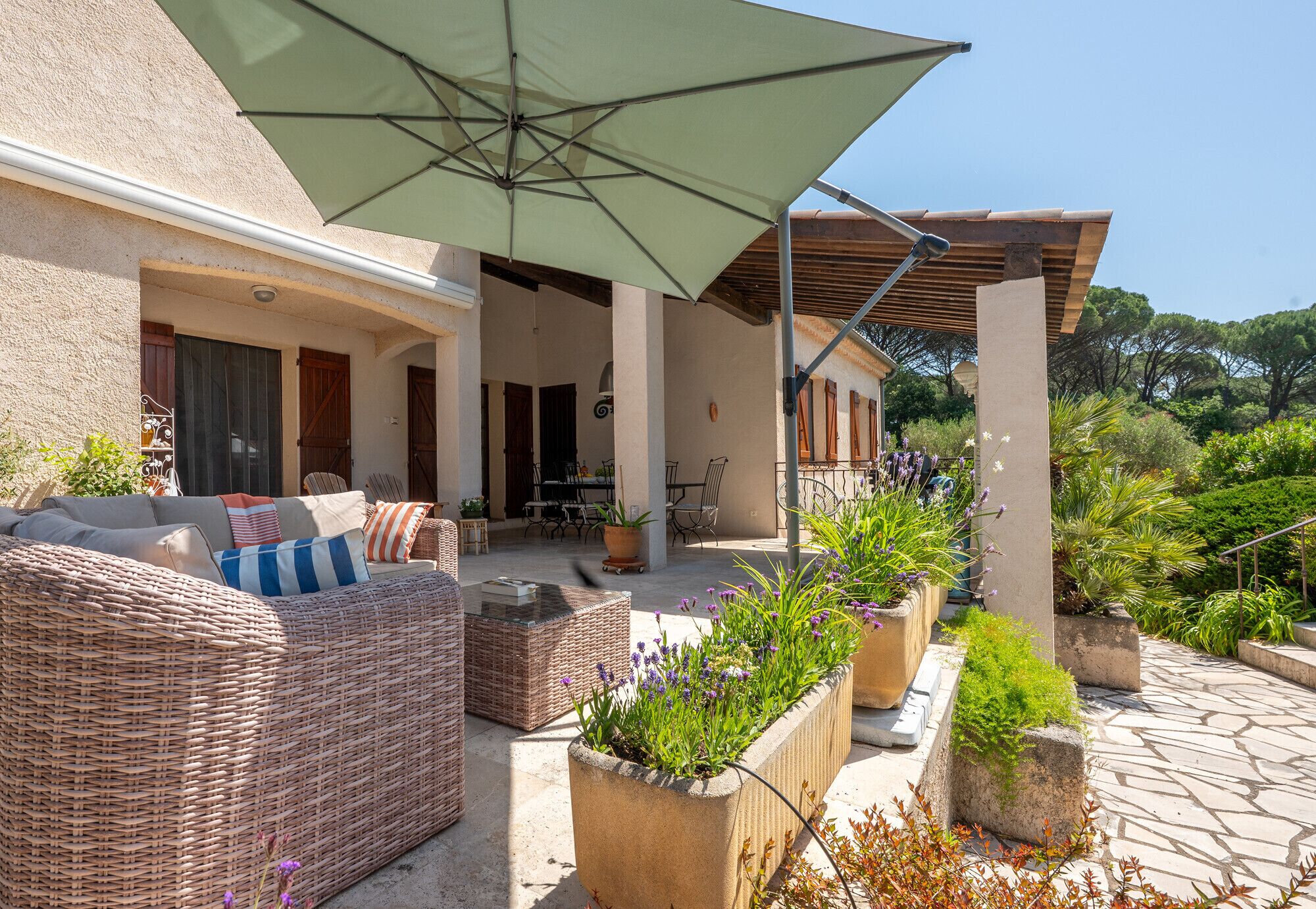 Een overdekt terras met rieten meubilair, plantenbakken met bloemen en een grote parasol.