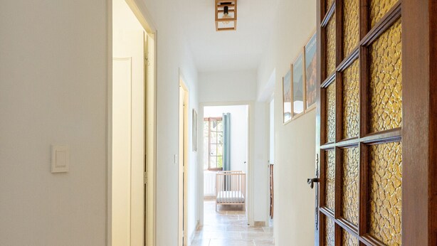 Um corredor com uma porta à esquerda, um luminário na parede e vista para uma varanda com guarda-corrimão.