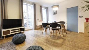 A modern living room with a flat-screen TV, a dining area with chairs, and a large window with curtains. - Spacious, recently renovated apartment near the Louvre (Île-de-France)