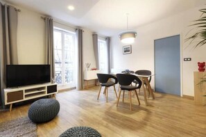 Ein modernes Wohnzimmer mit Flachbildfernseher, einem Essbereich mit Stühlen und einem großen Fenster mit Vorhängen.