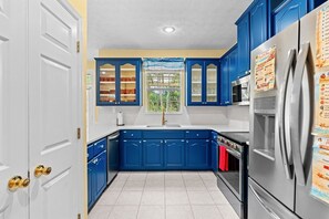 A kitchen with blue cabinets, a stainless steel refrigerator, and a window with a view of greenery. - The Love Bug: Private Pool, Golf Cart & Fire Pit, Sleeps 16 & Pet Friendly (Panama City Beach)