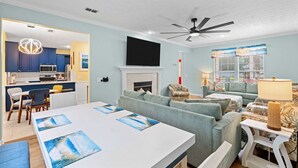 Ein modernes Wohnzimmer mit Kamin, einem Essbereich mit weißem Tisch und einer Küchenzeile mit blauen Schränken und einer Pendelleuchte.