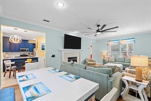 A modern living room with a fireplace, a dining area with a white table, and a kitchen area with blue cabinets and a pendant light. - The Love Bug: Private Pool, Golf Cart & Fire Pit, Sleeps 16 & Pet Friendly (Panama City Beach)