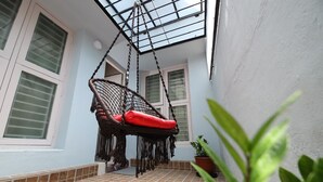 Una cadira penjant amb coixí vermell en una zona exterior coberta, amb sòl de rajola i plantes en testos.
