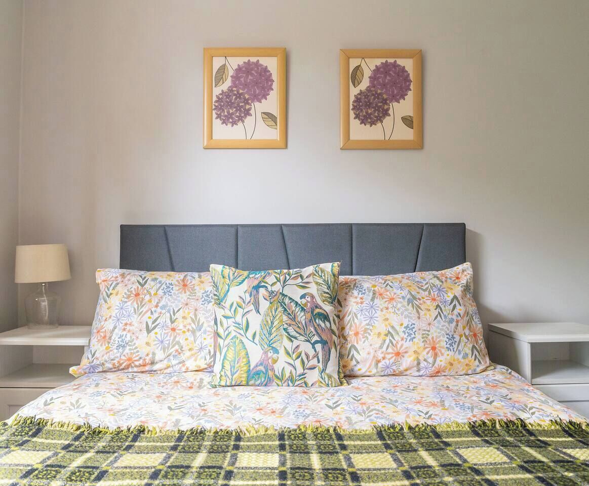 A bedroom with a bed, two pillows with floral patterns, a headboard, and two wall-mounted pictures of purple flowers.