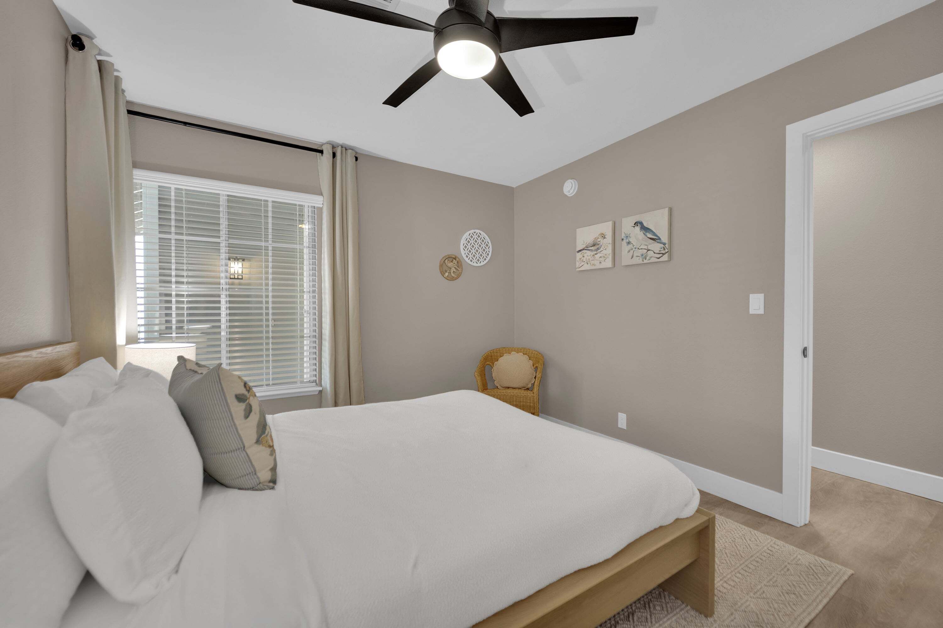 A bedroom with a bed, a ceiling fan, a window with blinds, a chair, and wall art.