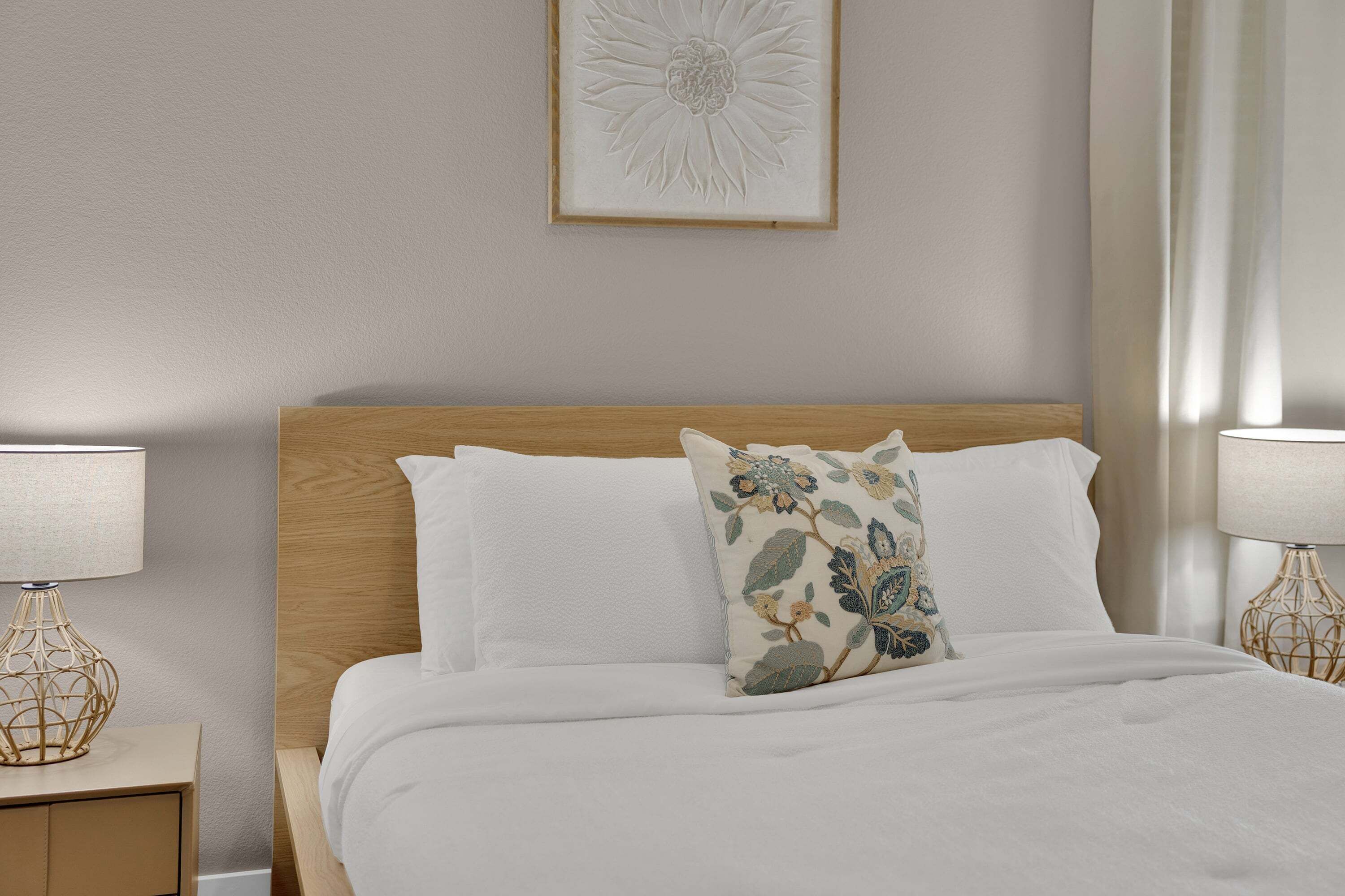 A neatly made bed with white linens and a floral pillow, flanked by two bedside tables with lamps, in a room with a simple, clean design.