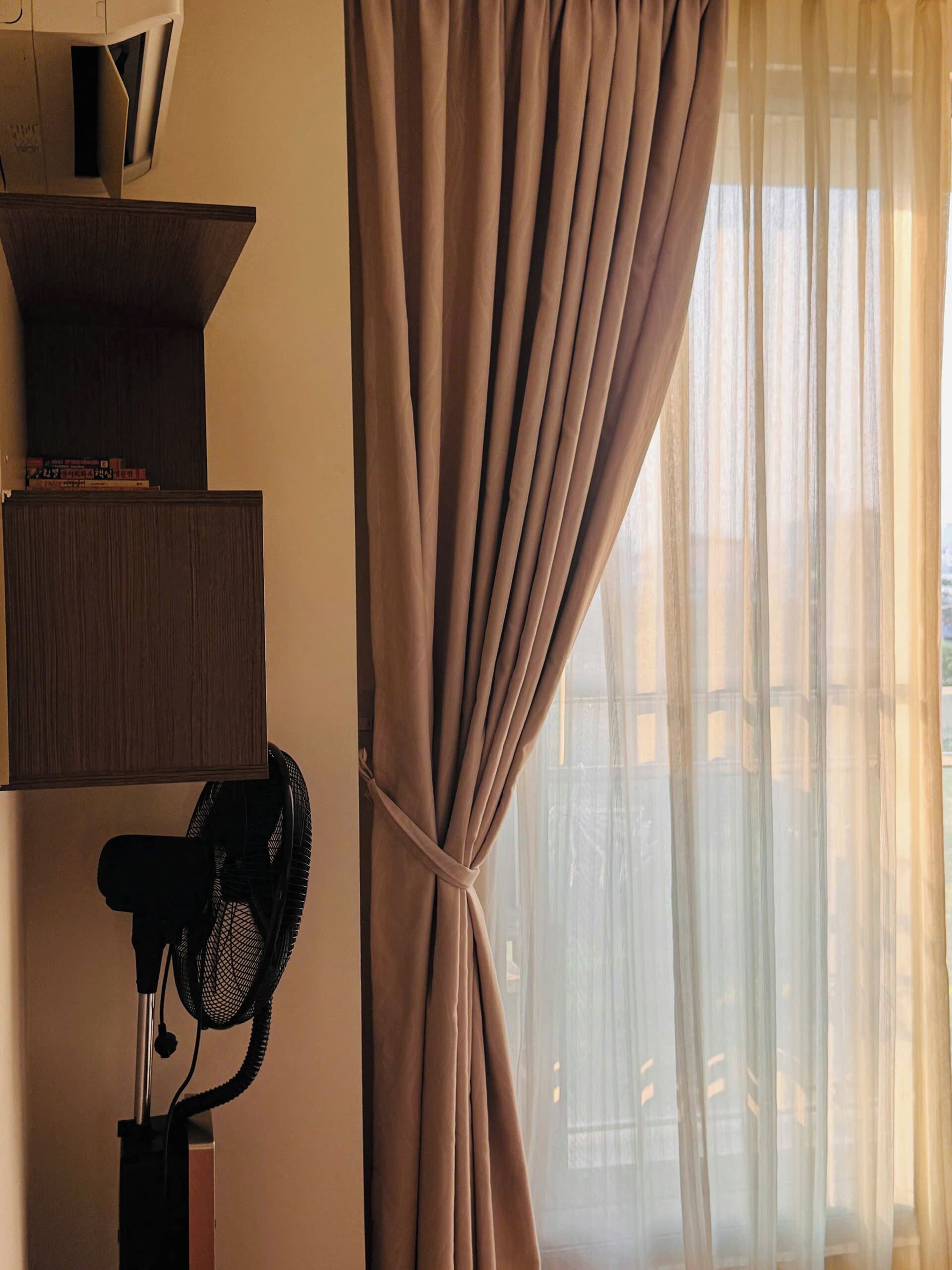 A room with a standing fan, a bookshelf, and a window with sheer curtains.