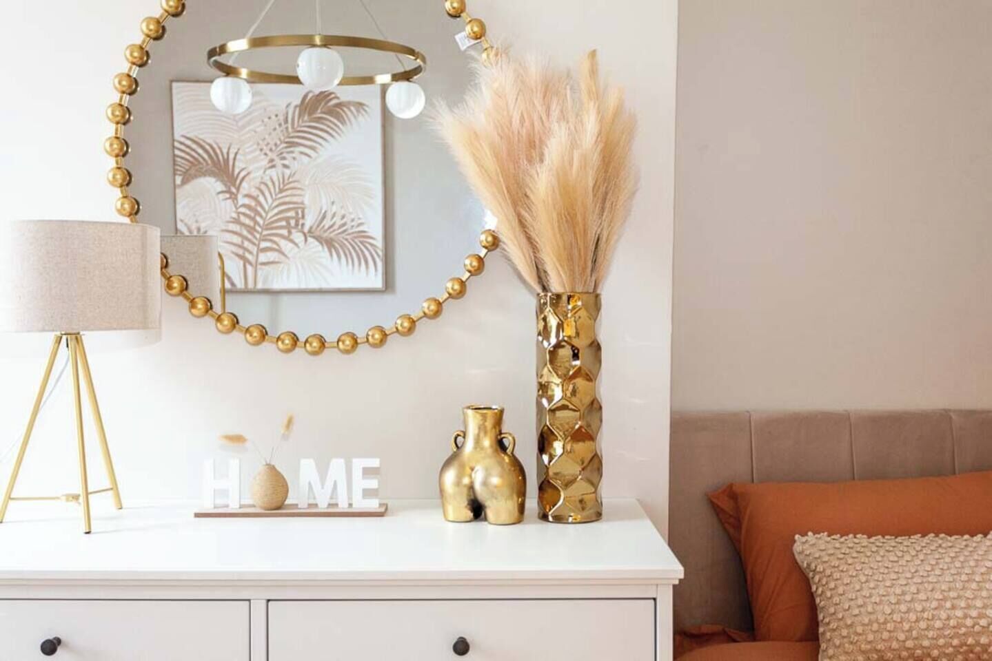 Une commode blanche, une lampe dorée, un vase doré et une œuvre d’art murale.