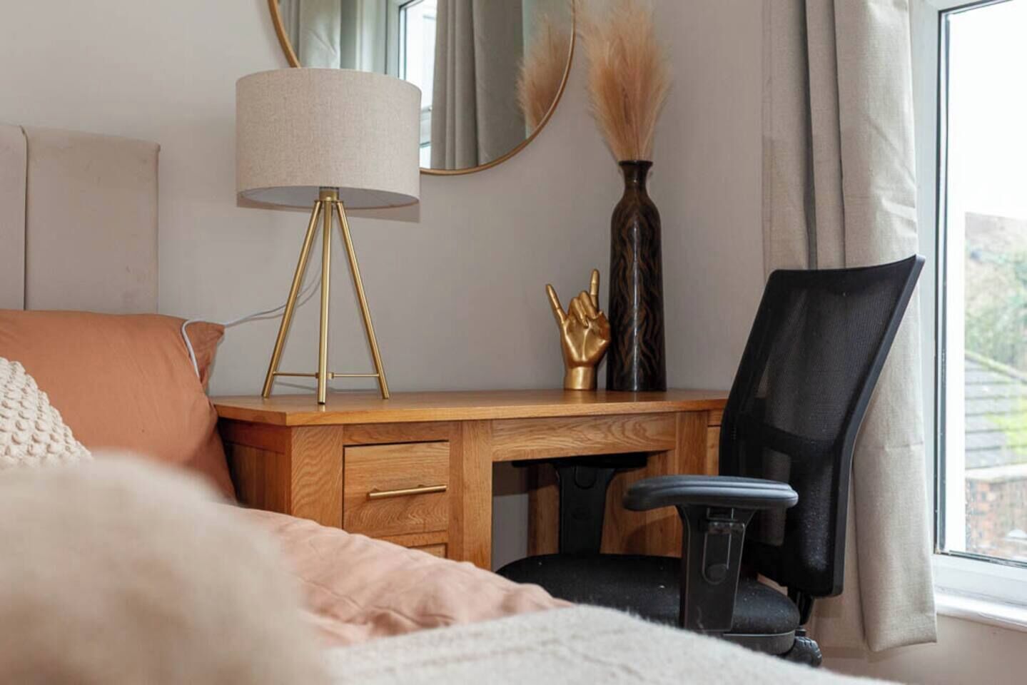Ein Schlafzimmer mit Schreibtisch aus Holz, einem schwarzen Bürostuhl, einer Lampe, einer Vase mit Pampasgras und einem Bett mit weißer Decke.