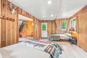 Una habitación con paredes de madera, una cama con colcha estampada, un escritorio con lámpara y una puerta que da a vistas de vegetación.