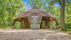 Una estructura rústica de madera con techo inclinado, una puerta y una ventana, rodeada de árboles.