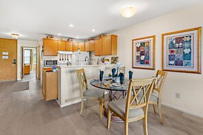 A modern kitchen with wooden cabinets, a central island, and a dining area with a glass table and chairs. - Hawaiian Getaway | 1BR Walk to Beach (Kihei)