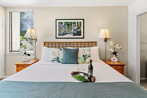 A hotel room with a bed, two bedside lamps, a framed picture on the wall, and a window with blinds. - Hawaiian Getaway | 1BR Walk to Beach (Kihei)