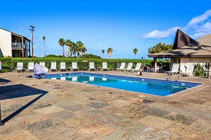 A swimming pool with lounge chairs and a covered seating area, surrounded by palm trees and a building. - Hawaiian Getaway | 1BR Walk to Beach (Kihei)