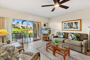 A living room with a sofa, a coffee table, and a TV. There is a view of a pool and palm trees outside. - Hawaiian Getaway | 1BR Walk to Beach (Kihei)