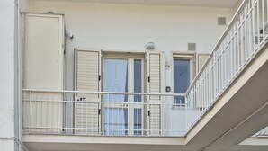 Uma varanda com guarda-corpo branco, uma porta com persianas e uma janela com cortina.