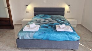 Uma cama com colcha floral, duas toalhas brancas, uma mesa de cabeceira e um aparelho de luz fixado na parede.