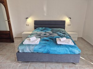 Ein Bett mit floralem Überwurf, zwei weiße Handtücher, ein Nachttisch und eine Wandleuchte.